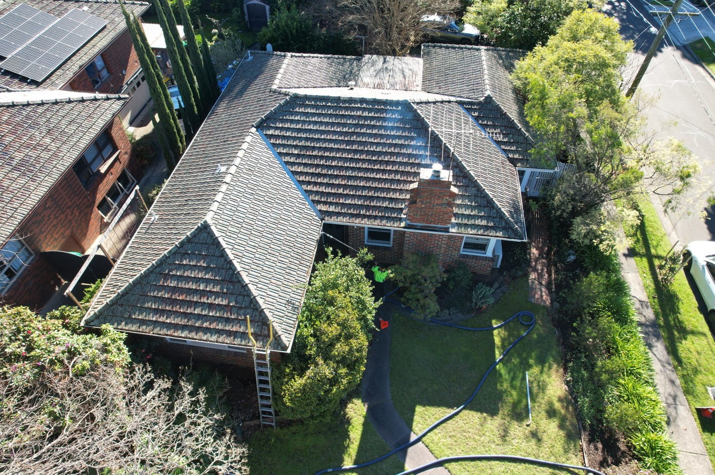 Aerial view of a moss covered tile roof with a ladder set against the gutter and vacuum hose on the lawn